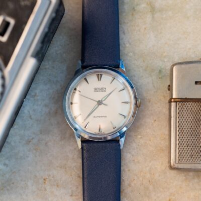 A vintage Gruen Precision Autowind watch with a silver dial and dark blue leather strap, placed between a camera and a metal lighter, available for sale.