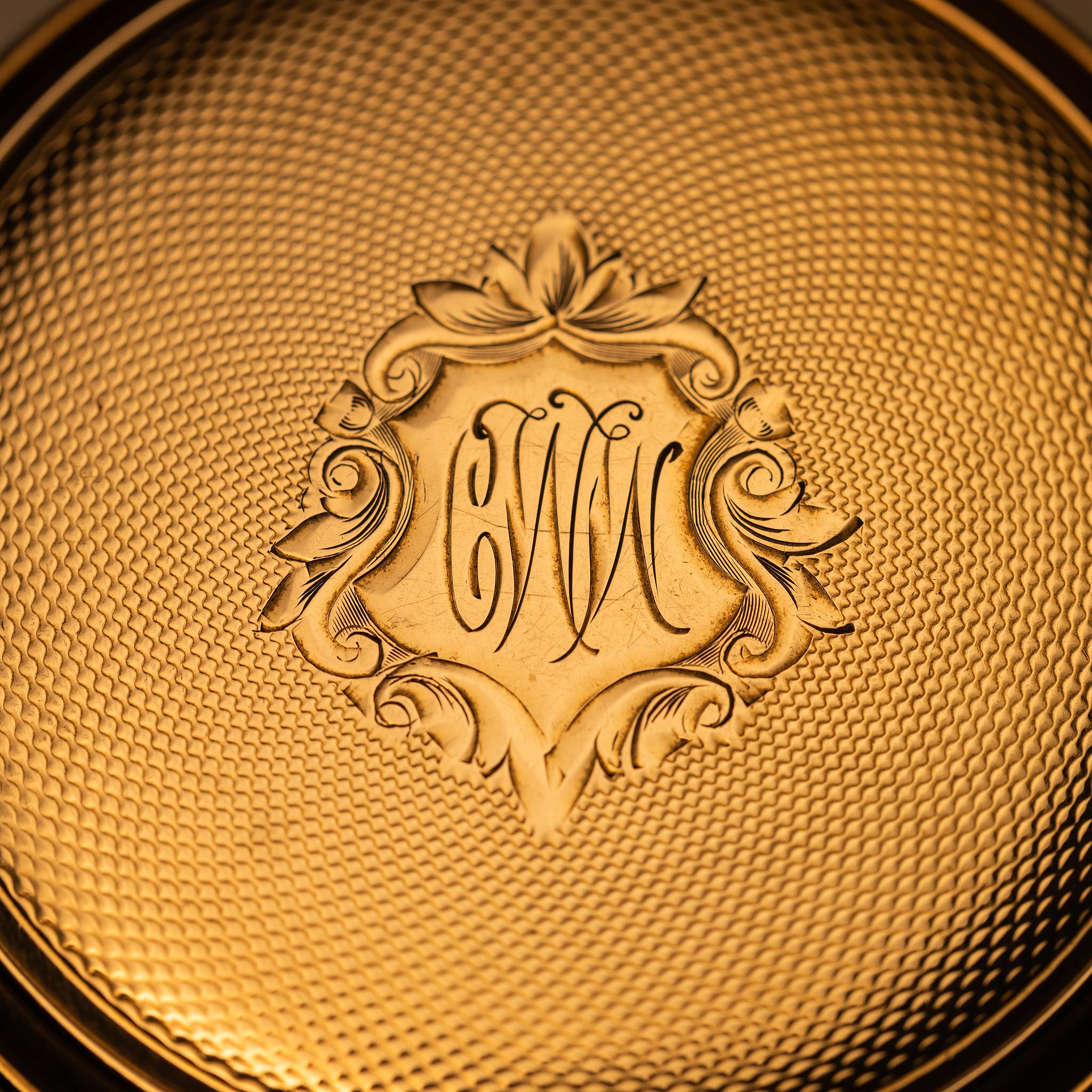 Close-up of a gold, textured surface featuring an ornate, engraved floral crest with the intertwined initials CMC in the center, evoking vintage elegance ideal for Rockford Watch Co. collectors or resale enthusiasts.