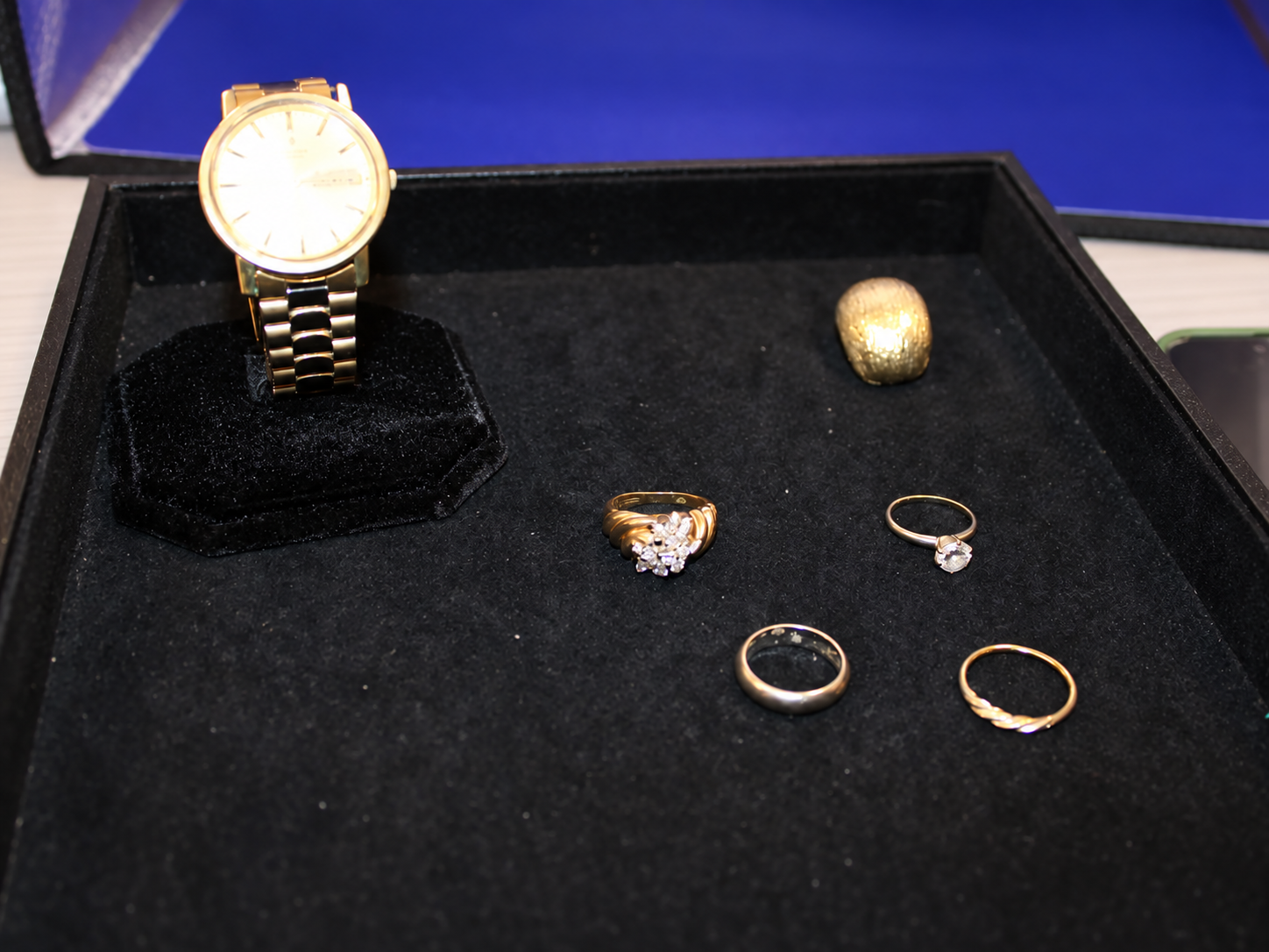 Actual safe-deposit-box auction lot photograph with an indistinct gold wristwatch, rings and a gold-colored piece of jewelry arranged in a black display tray